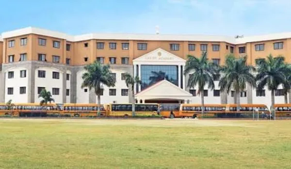 Christ Academy ICSE School, Bangalore — long ochre school building with a gabled entrance porch and a line of school buses drawn up in front of the playing field
