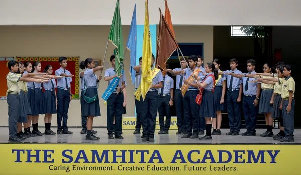 The Samhita Academy, Bangalore — pupils in uniform holding house flags on a stage above the school's name board